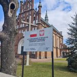 _m3ieUnc Kościół neogotycki z czerwoną cegłą w tle. Na pierwszym planie drewniany krzyż i tablica informacyjna. Obok drzewo, niebieskie niebo.