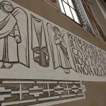 tQOYB2cs Mur z jasnym freskiem z postaciami aniołów obok napisu w języku polskim. Tło beżowe z ozdobnym białym wzorem. Obok są witraże, wpuszczające światło do wnętrza.
