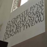 tdTuPV56 Ściana z czarnym napisem w kościele, obok okno wpuszcza światło. Po lewej złote detale na jasnym tle.