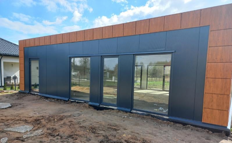 Nowoczesny budynek o fasadzie w odcieniach szarości i drewna. Duże szklane okna. W tle błękitne niebo i trawa.