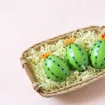 Koszyk z trzema zielonymi jajkami przypominającymi kaktusy wypełniony jest białymi wiórkami. Na jajkach są czarne krzyże. Tło jest jasnoróżowe.