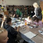 32 Dzieci malują przy długim stole w bibliotece, otoczeni książkami. Na stole są kolorowe pisaki i dekoracje. Tło wypełniają półki z książkami i jasne ściany.