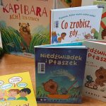 Stos książek dziecięcych w różnych kolorowych okładkach na drewnianym stole, ozdobiony figurką żaby.