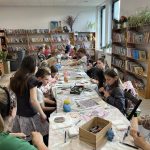 42 Grupa dzieci siedzi przy długim stole w bibliotece, malując kamienie. Otacza ich regał z kolorowymi książkami. Pokój jest jasny i przytulny.