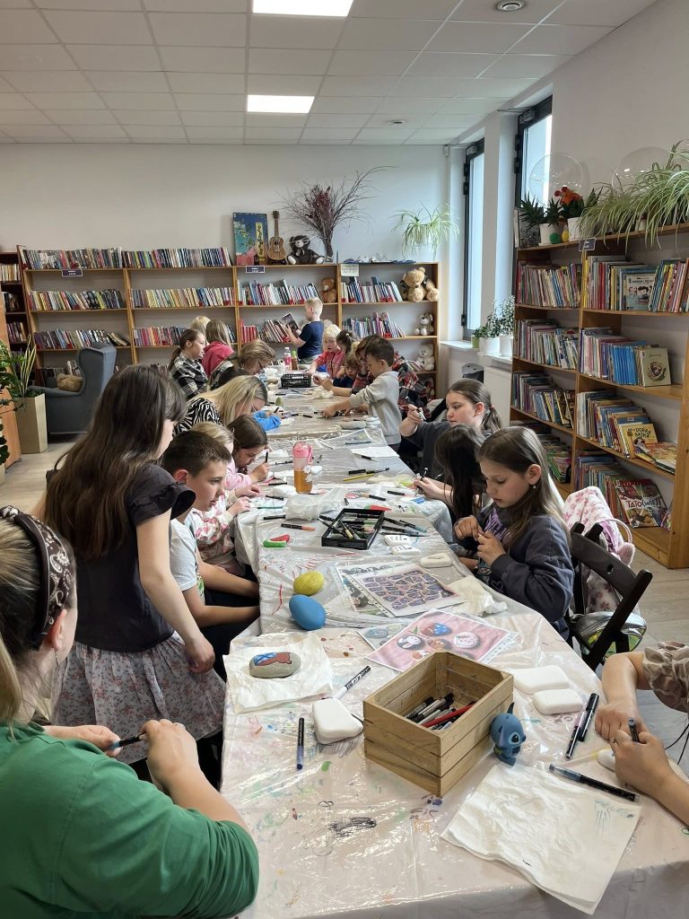 Grupa dzieci siedzi przy długim stole w bibliotece, malując kamienie. Otacza ich regał z kolorowymi książkami. Pokój jest jasny i przytulny.