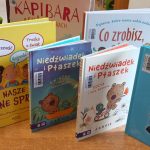 Na zdjęciu widoczne są kolorowe książki dla dzieci na drewnianej półce. Okładki w jasnych barwach, z dużymi, kolorowymi napisami i ilustracjami. W tle znajduje się zielona dekoracja w kształcie lisa.