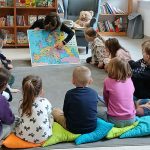 Grupa dzieci siedzi na poduszkach wokół nauczyciela, który pokazuje mapę Europy. W tle widoczne są kolorowe książki na półkach. Pomieszczenie jest jasne i przestronne.