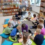 Zdjęcie przedstawia dzieci siedzące na kolorowych poduszkach w bibliotece. Mają książki na szarym dywanie. W tle są półki z książkami i rośliny.