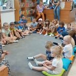 Grupa dzieci siedzi w kręgu na kolorowych poduszkach w bibliotece. Na drewnianych regałach w tle widać mnóstwo książek. Dwie nauczycielki są w pobliżu, jeden w brązowej sukni siedzi na ziemi z dziećmi