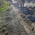 Pracownicy naprawiają gruntową drogę, podnosząc kłęby kurzu. Obok zielone drzewa i dom z białym płotem. Jasne, słoneczne popołudnie.