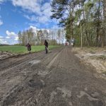 Dwóch ludzi pracuje na polnej drodze, otoczonej drzewami, przy jasnym niebie. Jeden trzyma łopatę, obok pole i las. W tle czerwony pojazd.