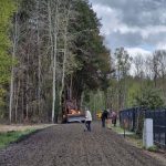 Na zdjęciu widoczny jest traktor na drodze otoczonej drzewami, pracujący nad utwardzaniem powierzchni. Tło to zieleń lasu i szare niebo z chmurami. Kilku ludzi z narzędziami pochyla się nad ziemią.