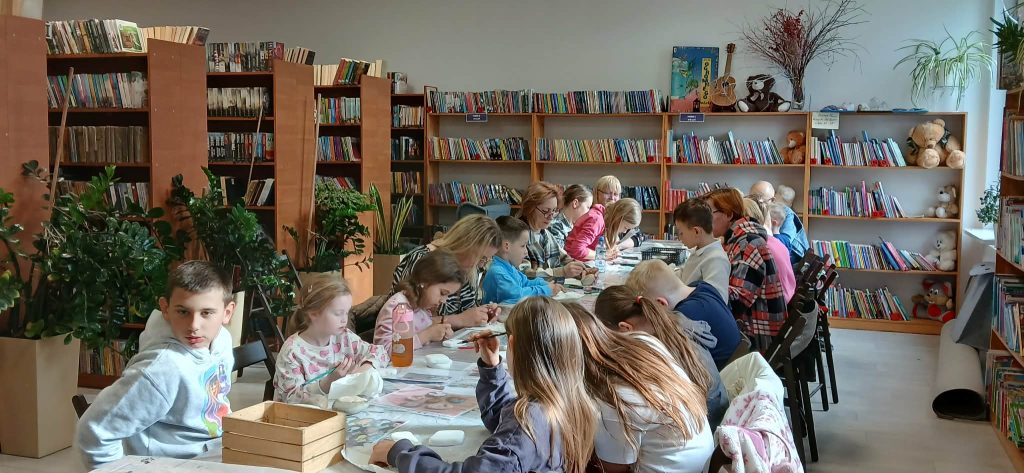 Grupa dzieci maluje w bibliotece. W tle półki pełne kolorowych książek i rośliny. Atmosfera jest jasna i kreatywna.