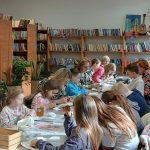 Grupa dzieci maluje w bibliotece. W tle półki pełne kolorowych książek i rośliny. Atmosfera jest jasna i kreatywna.