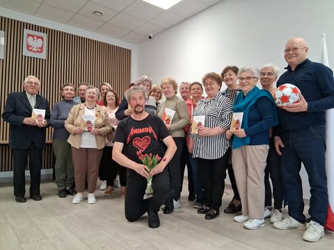 Grupa ludzi w różnym wieku stoi wewnątrz jasnego pokoju. Mężczyzna w koszulce z sercem klęczy z kwiatami. Na ścianie wisi emblemat z orłem.