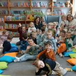 Grupa dzieci siedzi na podłodze w bibliotece, otoczona kolorowymi poduszkami. W tle regały z książkami. Dwoje dorosłych pośród dzieci.