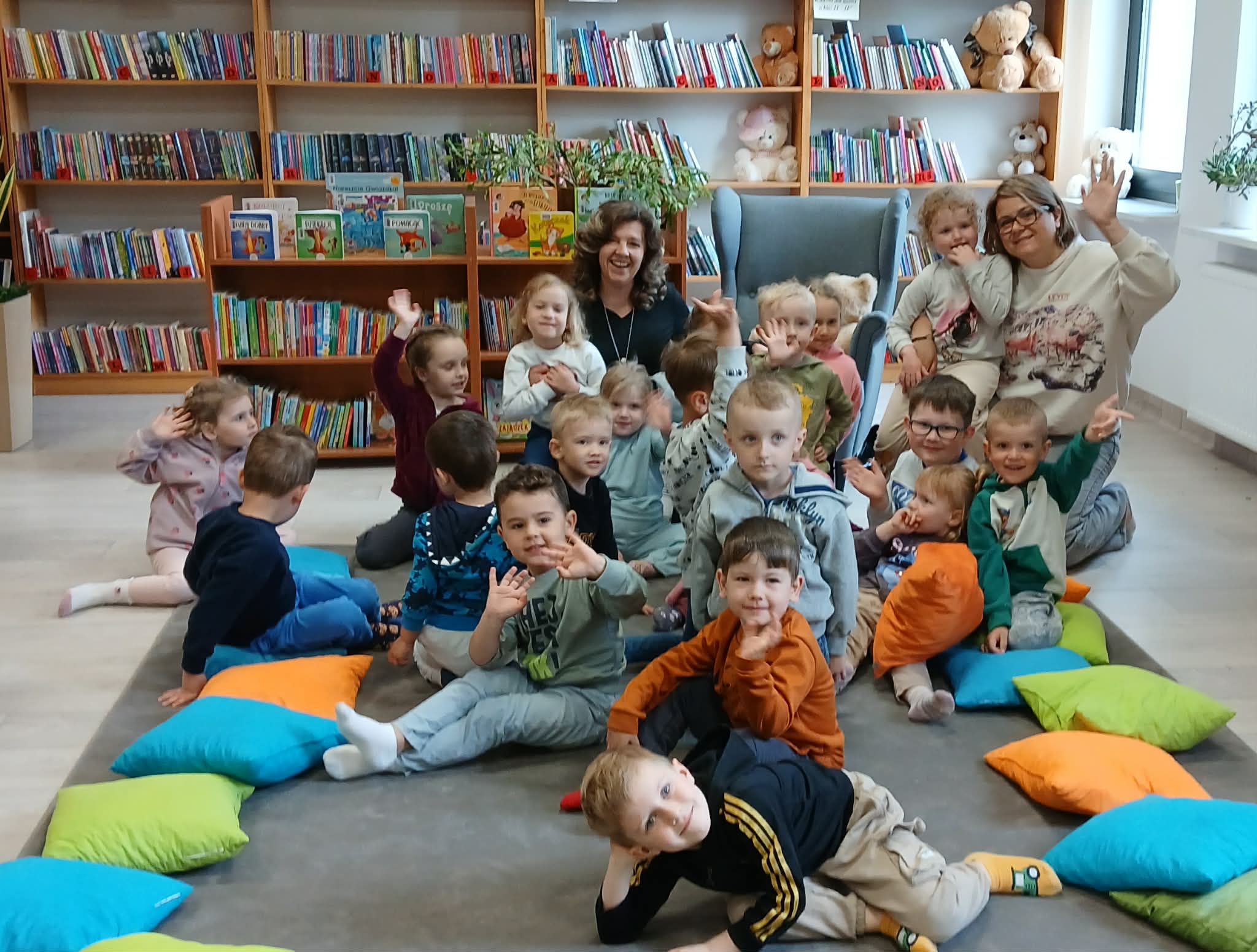 Grupa dzieci siedzi na podłodze w bibliotece, otoczona kolorowymi poduszkami. W tle regały z książkami. Dwoje dorosłych pośród dzieci.