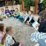 Dzieci siedzą w kręgu na kolorowych poduszkach, kobieta czyta im książkę. W tle widać regały z książkami i rośliny. Klimat biblioteki jest przyjazny i ciepły.