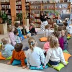 Grupa dzieci słucha kobiety w bibliotece, siedząc na kolorowych poduszkach. Tło: półki z książkami i rośliny. W prawym rogu stoi fotel z misiem.