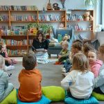 Grupa dzieci siedzi na kolorowych poduszkach w czytelni, słuchając kobiety czytającej książkę. Otoczeni są półkami z książkami i roślinami. W tle widać jasne ściany i duże okno.