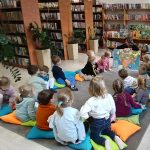 Grupa dzieci siedzi na kolorowych poduszkach w bibliotece. Kobieta pokazuje im mapę. W tle są półki z książkami i rośliny.
