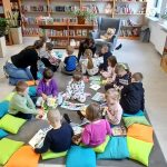 Dzieci siedzą na kolorowych poduszkach w bibliotece, otoczone półkami z książkami. Tło jest jasne, z roślinami i oknami po prawej. W tle regały pełne różnorodnych książek.