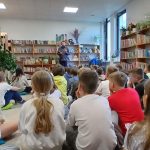Grupa dzieci siedzi na podłodze w jasnej bibliotece. U góry pełne książki regały, a w tle osoba czyta. Rośliny ozdabiają wnętrze.