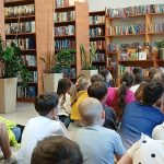 Dzieci siedzą na podłodze w bibliotece, słuchając mężczyzny z książką. Za nim regały pełne książek, zielone rośliny. Kolory drewniane, jasne i zielone.