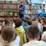 Grupa dzieci siedzi na podłodze w bibliotece z książkami na półkach. Dorosły mężczyzna czyta na tle pełnym kolorowych książek. Wokół rośliny i jasne światło z okna.