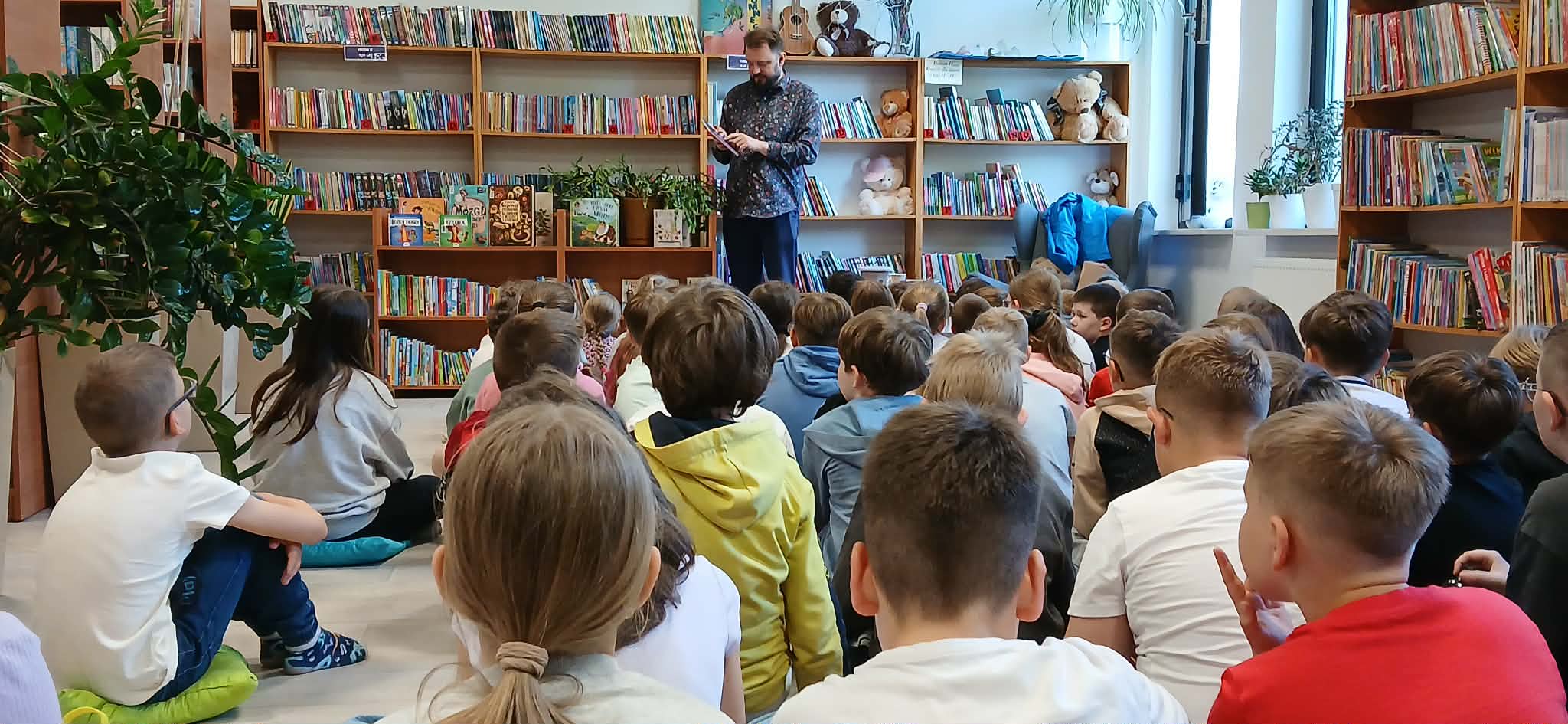 Grupa dzieci siedzi na podłodze w bibliotece z książkami na półkach. Dorosły mężczyzna czyta na tle pełnym kolorowych książek. Wokół rośliny i jasne światło z okna.