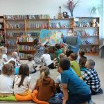 Na zdjęciu widać dzieci siedzące na poduszkach w bibliotece. Przed nimi osoba pokazuje mapę. W tle są kolorowe regały z książkami i rośliny.