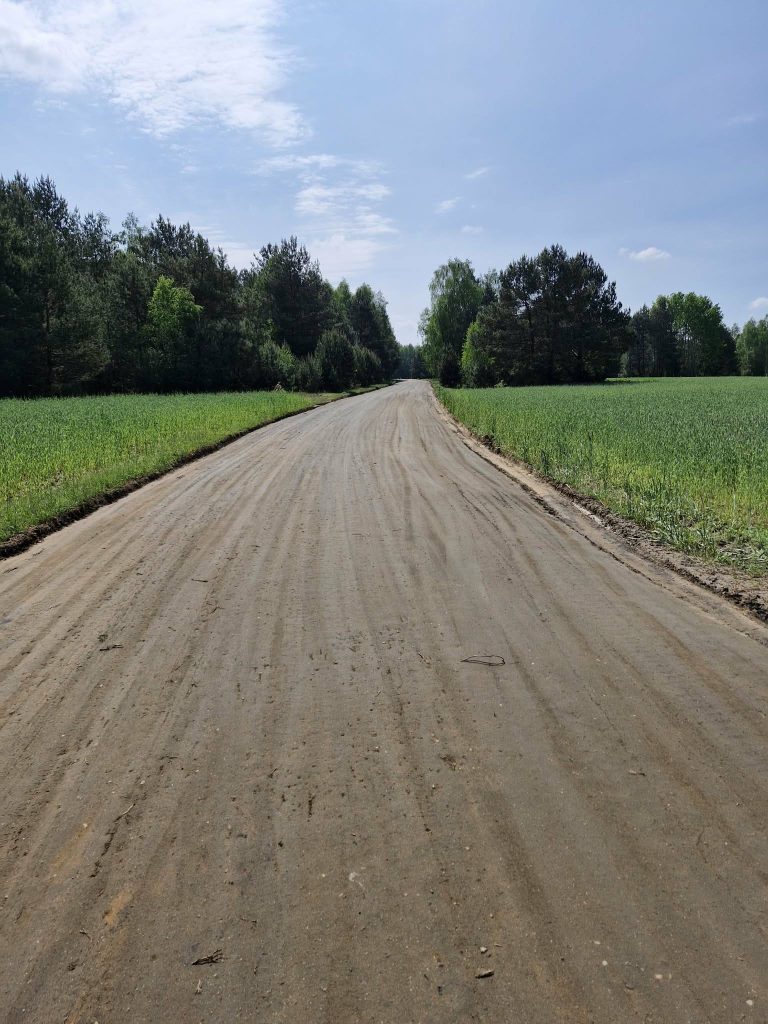 Szeroka, piaszczysta droga prowadzi przez zielone pola z lasem w tle. Drzewa są gęste, niebo jest jasnoniebieskie z chmurami. Droga biegnie na horyzoncie, tworząc spokojny pejzaż.