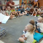 Dzieci siedzą na poduszkach w kole, słuchając czytanej książki. Kobieta z książką ma sukienkę w niebieskie wzory. W tle regały z kolorowymi książkami i inne osoby.