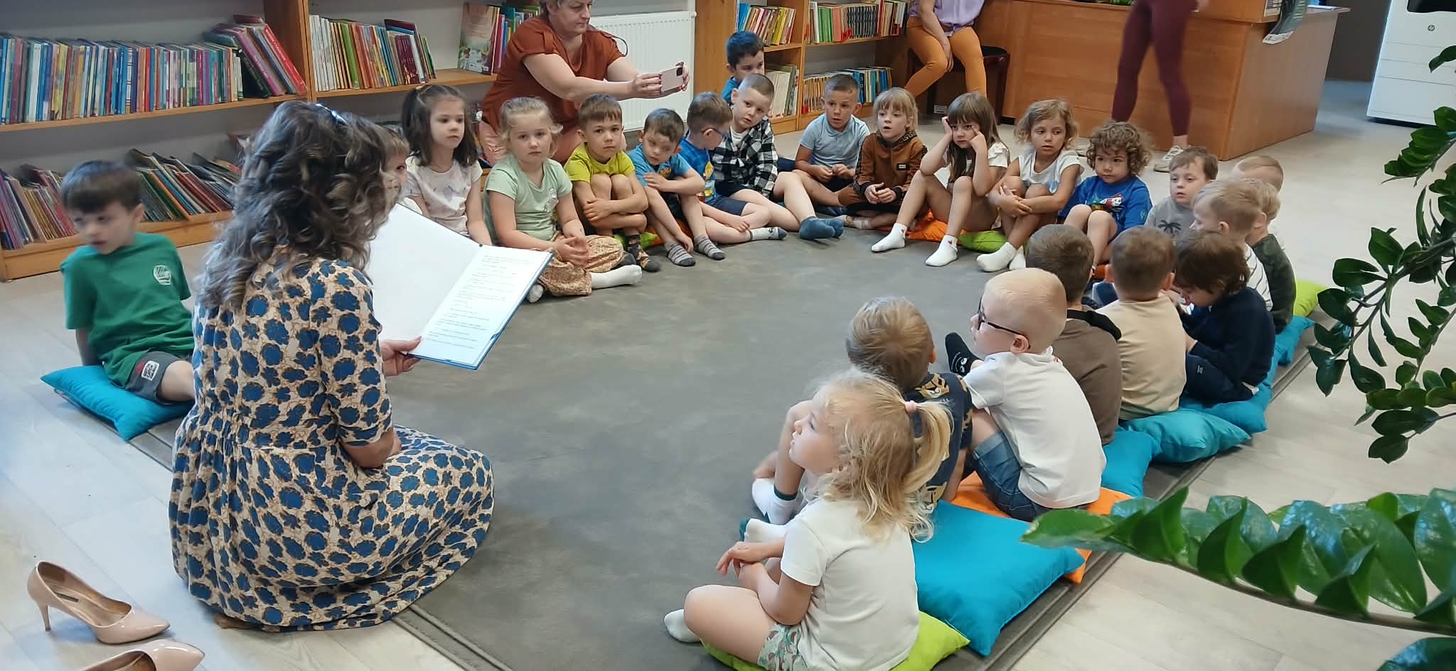Dzieci siedzą na poduszkach w kole, słuchając czytanej książki. Kobieta z książką ma sukienkę w niebieskie wzory. W tle regały z kolorowymi książkami i inne osoby.