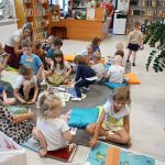 Grupa dzieci siedzi na poduszkach na podłodze w bibliotece pełnej kolorowych książek. Dorośli stoją i pomagają. W tle regały z książkami i rośliny.