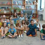 Grupa dzieci siedzi na dywanie w bibliotece. W tle regały z kolorowymi książkami. Obok dzieci siedzi kobieta.