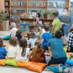 Grupa dzieci siedzi na kolorowych poduszkach w bibliotece. Kobieta pokazuje im książkę. W tle widoczne są regały pełne książek.