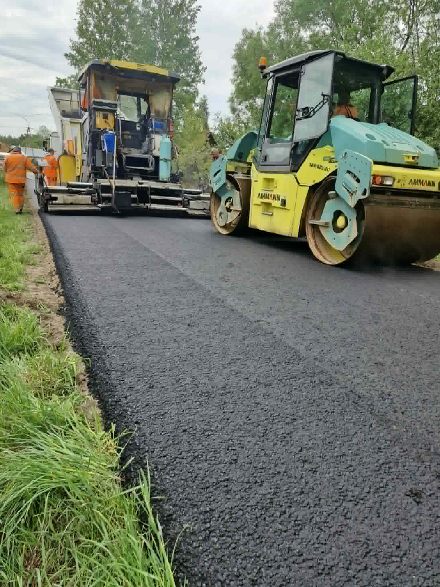Budowa drogi z nowym asfaltem. Widoczny walec i maszyna do asfaltu w jasnozielonym kolorze. W tle drzewa i pracownicy w pomarańczowych kombinezonach.