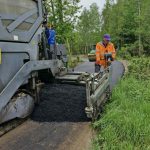Robotnik w pomarańczowym stroju obsługuje asfaltowanie drogi. Szara maszyna kładzie świeży asfalt. W tle zielone drzewa i trawa.