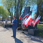 Mężczyzna w niebieskim garniturze stoi przed pomnikiem otoczonym polskimi flagami. Dwie osoby w mundurach harcerskich stoją obok. Tło tworzą drzewa i jasne, słoneczne niebo.