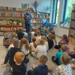 Pokój z półkami pełnymi książek, dzieci siedzą na podłodze. Mężczyzna czyta książkę, w tle duże okna i rośliny. Atmosfera edukacyjna.