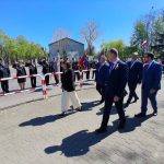 Grupa osób w eleganckich strojach idzie chodnikiem. W tle znajdują się strażacy w mundurach i flagi. Jest słoneczny dzień, a niebo jest czyste.