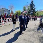 Na zdjęciu widoczni są ludzie w eleganckich strojach w otoczeniu pocztów sztandarowych. Tło stanowią zielone drzewa i niebieskie niebo. Wydarzenie odbywa się na zewnątrz, na brukowanej powierzchni.
