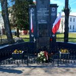 Pomnik z tablicą upamiętniającą, otoczony jest flagami Polski i kwiatami. Dwie harcerki w mundurach stoją na baczność. Tło stanowi ogrodzenie, drzewa i biały budynek.