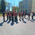 Orkiestra w czerwonych strojach gra na placu. Tło stanowi żółty budynek i drzewa pod jasnym niebem. Cień muzyków kładzie się na chodniku.