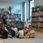 Mężczyzna czyta dzieciom w jasnej bibliotece. Dookoła są kolorowe półki z książkami. Na dywanie siedzi grupa dzieci.