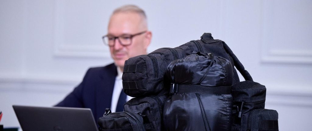 Zdjęcie przedstawia mężczyznę przy komputerze siedzącego na tle jasnej ściany. Na pierwszym planie widoczny jest czarny plecak. Tło jest jasne, co kontrastuje z ciemnym plecakiem.