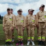 Grupa dzieci w beżowych uniformach z neonowymi akcentami stoi na trawie. Mają czapki z napisem STRAŻ, a w tle widać niebo z chmurami. Wszystko w jasnej tonacji na tle zieleni.