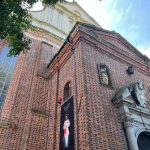 Zdjęcie przedstawia fasadę kościoła z czerwonej cegły. Na murze wisi baner z Jezusem Miłosiernym. W tle widać niebieskie niebo i drzewa.