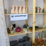 Drewniane półki z glinianymi figurkami i malarskimi przyborami na tle białej ściany. Dominują beże, biel i szarość. Na środkowej półce logo Wienerberger.